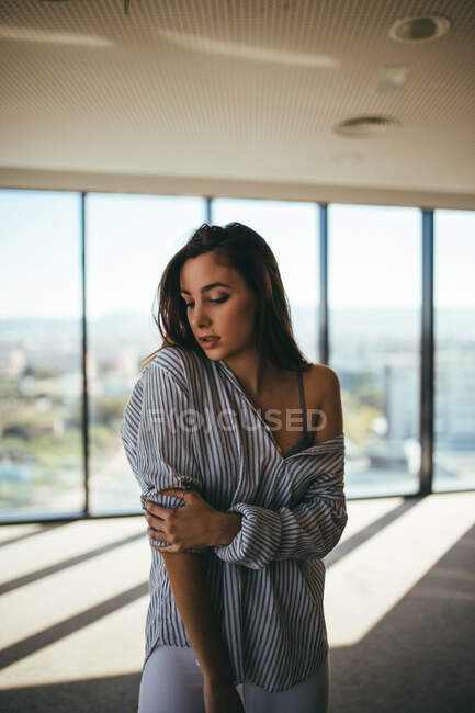 Back view of dreamy sensual beautiful woman in striped shirt showing naked shoulder while standing in room with panoramic windows with closed eyes — Stock Photo