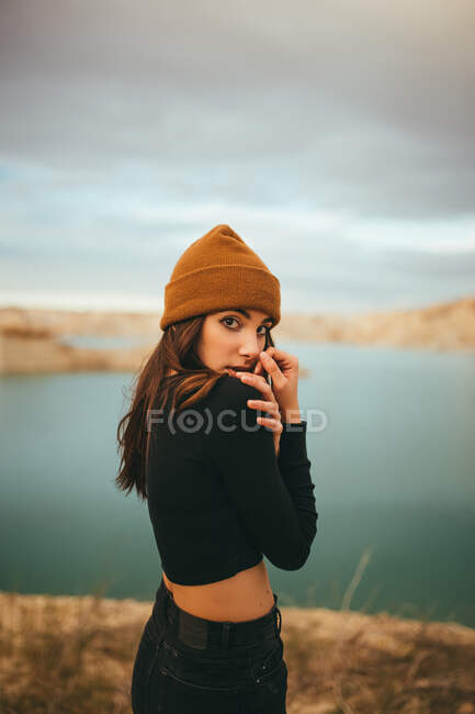 Romántica joven sensible linda mujer en ropa casual mirando a la cámara sobre el hombro mientras está de pie en la costa del lago en colorido atardecer - foto de stock
