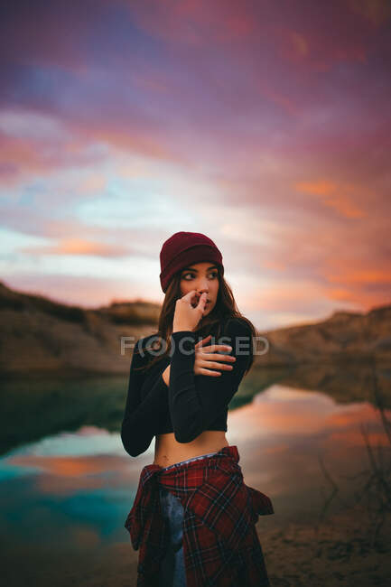 Romantique jeune femme mignonne sensible en vêtements décontractés regardant loin tout en se tenant sur la côte du lac au coucher du soleil coloré — Photo de stock