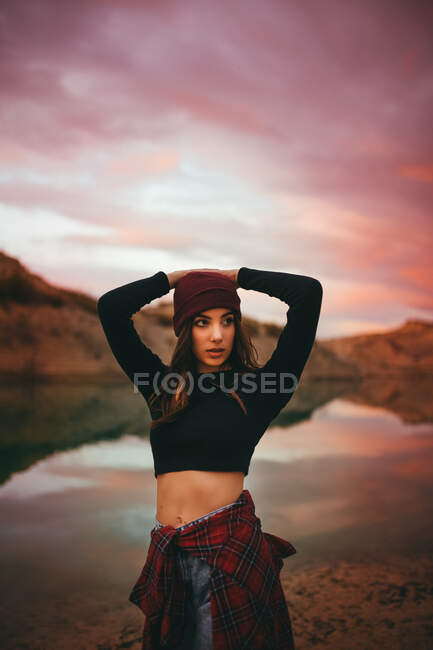 Romantico giovane donna sveglia sensibile in abiti casual guardando lontano e tenendosi per mano dietro la testa mentre in piedi sulla costa del lago in tramonto colorato — Foto stock