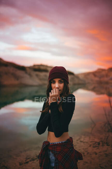Mulher bonito sensível juvenil romântico em roupas casuais olhando para a câmera enquanto estava na costa do lago em pôr do sol colorido — Fotografia de Stock