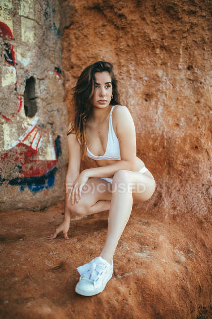 Знизу чуттєва молода чарівна жінка в білому бікіні і кросівки дивиться далеко, присідаючи на скелю — стокове фото
