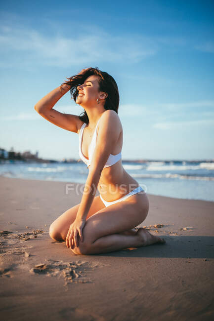 Вид сбоку веселой молодой туристки в купальниках, сидящей на коленях в солнечном свете на песчаном пляже во время летнего отпуска к морю — стоковое фото