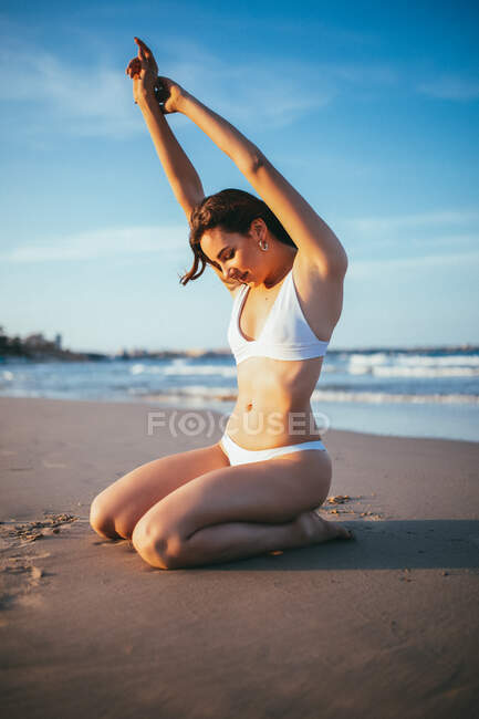 Vista laterale di gioiosa giovane turista donna in costume da bagno seduta sulle ginocchia alla luce del sole sulla spiaggia di sabbia durante le vacanze estive al mare — Foto stock