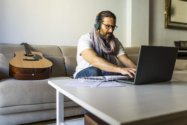 Male freelancer in casual wear and eyeglasses typing on keyboard of laptop in headphones while sitting on sofa — Stock Photo