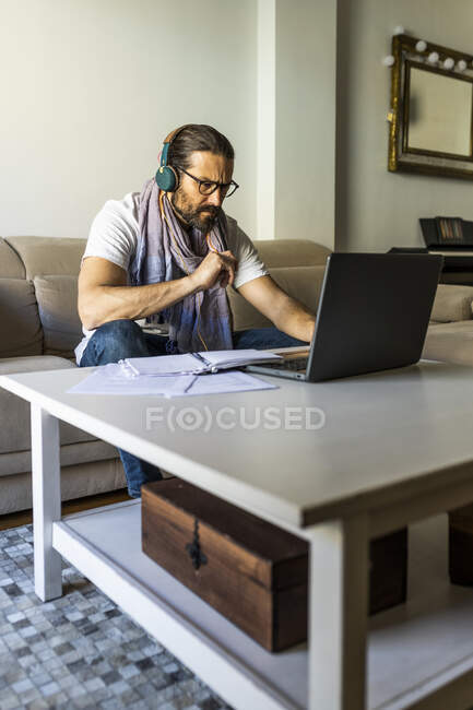 Homme barbu sérieux en tenue décontractée et lunettes regarder ordinateur portable tout en écoutant de la musique avec écouteurs dans le salon élégant — Photo de stock