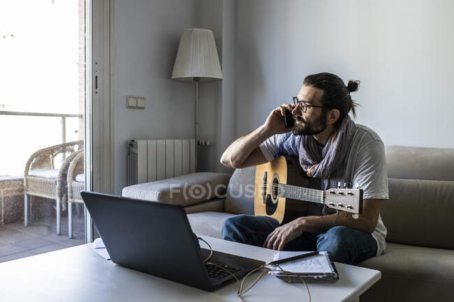 Творческий занятой мужчина-музыкант в повседневной одежде и модных очках сидит на диване с гитарой, используя ноутбук и разговаривая по мобильному телефону в гостиной — стоковое фото