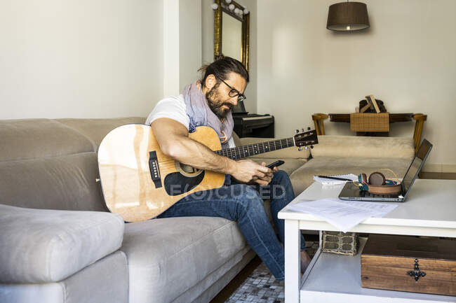 Fröhlicher Mann mit Gitarre checkt Smartphone auf Sofa — Stockfoto