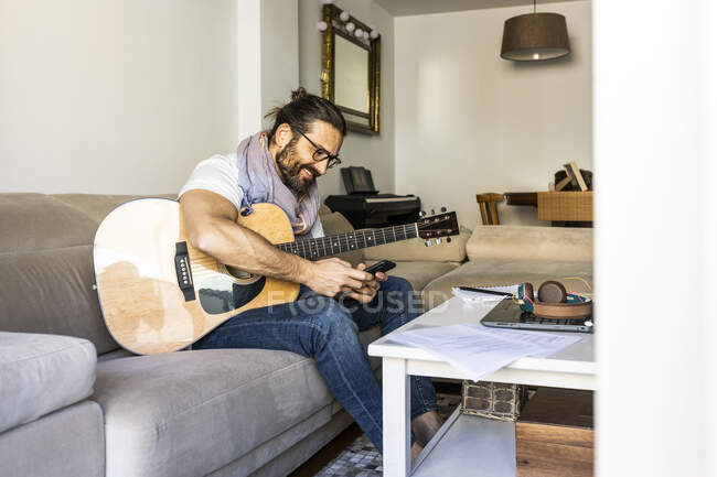 Homem alegre com guitarra verificando smartphone no sofá — Fotografia de Stock