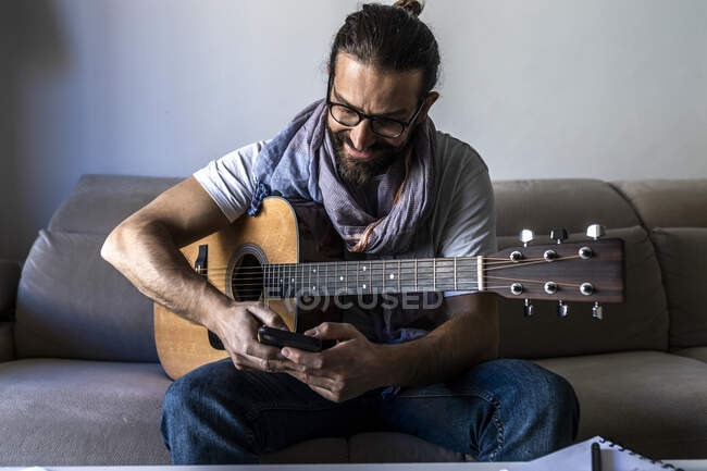 Контент-бородатий чоловічий музикант сидить на дивані з гітарою під час серфінгу на мобільному телефоні і посміхається в сучасній вітальні — стокове фото