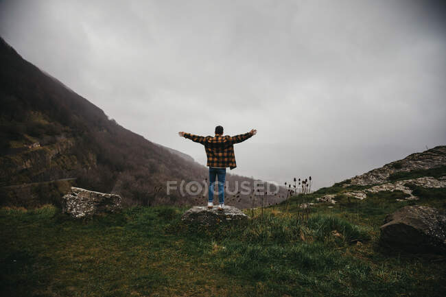 Dal basso vista posteriore dell'escursionista maschio irriconoscibile in abiti casual in piedi sulla scogliera e allargando le braccia in una giornata nuvolosa in campagna — Foto stock