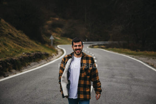 Der bärtige, schwarzhaarige, fröhliche Mann in lässiger Kleidung lächelt in die Kamera, während er auf einer leeren Straße in der herbstlichen Landschaft steht. — Stockfoto