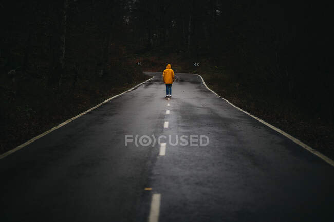 Visão traseira do homem anônimo em casaco amarelo andando na estrada de asfalto vazio entre a floresta verde — Fotografia de Stock