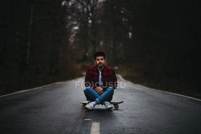 Unbekümmerter ethnischer Typ in stylischer Freizeitkleidung sitzt mit überkreuzten Beinen auf dem Skateboard auf asphaltierter Straße und blickt in die Kamera im herbstlichen Wald — Stockfoto