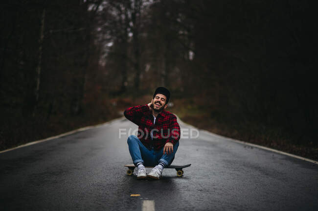 Беззаботный веселый мужчина в стильной повседневной одежде сидит со скрещенными ногами на скейтборде по асфальтовой дороге и смотрит в камеру среди осеннего леса — стоковое фото