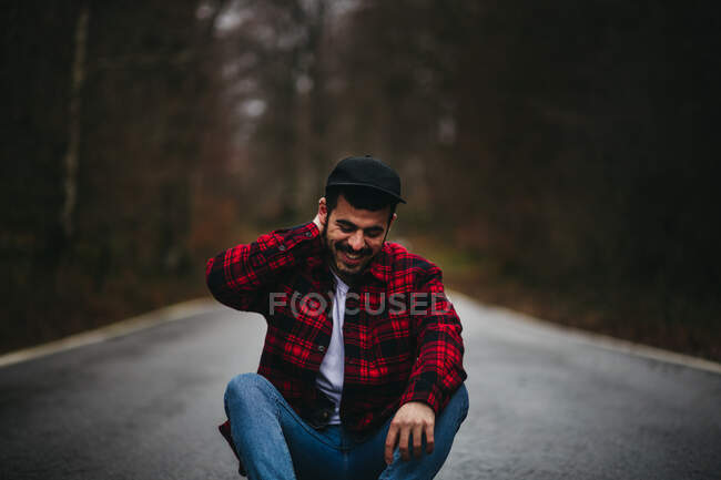 Беззаботный человек в стильной повседневной одежде сидит на асфальтированной дороге и смотрит вниз с закрытыми глазами среди осеннего леса — стоковое фото