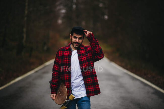Молодой стильный мужчина в повседневной одежде ходит по асфальтированной дороге со скейтбордом в руке осенним днем, глядя в камеру — стоковое фото