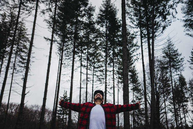 Young bearded male tourist in casual wear spreading arms and looking up among evergreen coniferous forest on overcast — Stock Photo