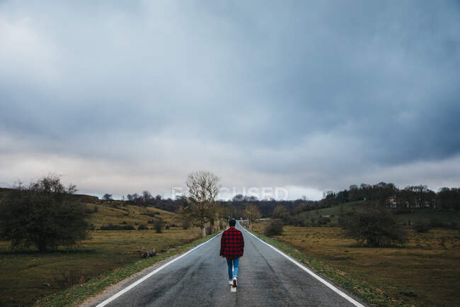 Back view of anonymous man in casual wear walking on empty asphalt road among green fields with cloudy sky on background — Stock Photo