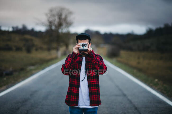 Неузнаваемый мужчина в стильной повседневной одежде, держащий фотокамеру перед лицом, стоя на дороге в сельской местности — стоковое фото