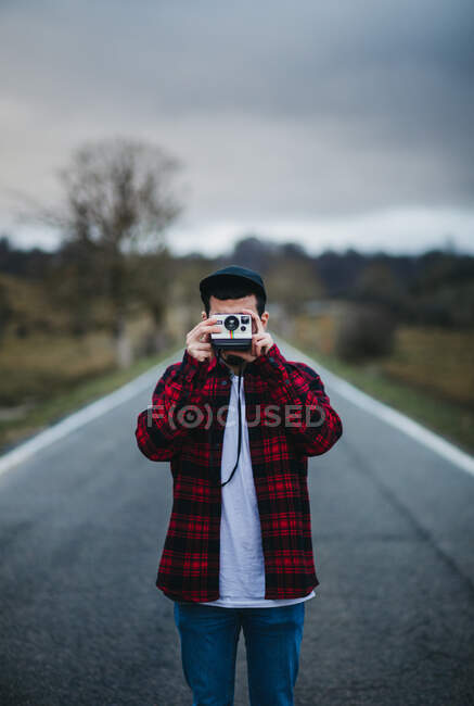 Невпізнаваний чоловік у стильному повсякденному вбранні тримає фотоапарат перед обличчям, стоячи на дорозі в сільській місцевості — стокове фото
