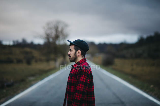 Vista lateral del hombre en ropa casual caminando por un camino de asfalto vacío entre campos verdes con cielo nublado en el fondo - foto de stock