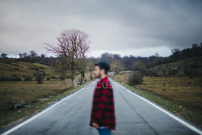 Vista laterale di anonimo uomo sfocato in abbigliamento casual che cammina su strada asfaltata vuota tra campi verdi con cielo nuvoloso sullo sfondo — Foto stock
