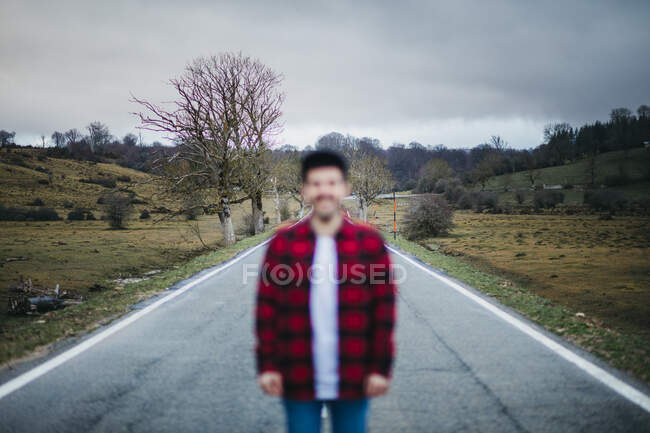 Hombre desenfocado anónimo en ropa casual caminando por un camino de asfalto vacío entre campos verdes con cielo nublado en el fondo - foto de stock