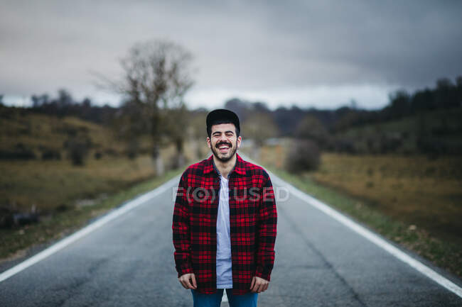 Cheerful man with closed eyes in casual wear standing on empty asphalt road among green fields with cloudy sky on background — Stock Photo