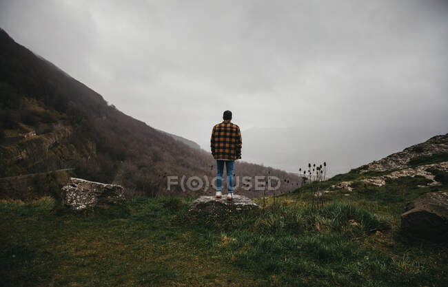 De baixo vista traseira de caminhante masculino irreconhecível em roupas casuais em pé no penhasco em dia nublado no campo — Fotografia de Stock