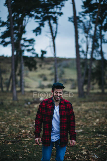 Hombre alegre en ropa casual de pie entre árboles de coníferas de hoja perenne en el campo de otoño - foto de stock