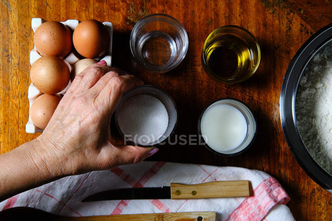 С вышесказанного анонимная женщина берет чашку сахара из деревянного стола рядом с различными ингредиентами во время приготовления пончиков на дому — стоковое фото