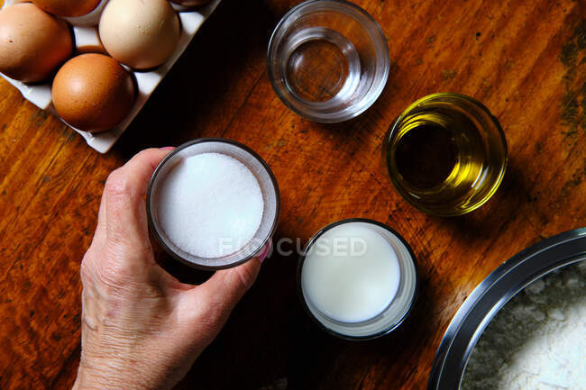 Зверху анонімна жінка бере чашку цукру з дерев'яного столу біля різних інгредієнтів під час приготування пончиків вдома — стокове фото