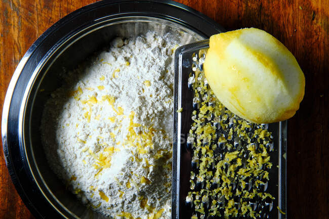 Vue de dessus de la râpe fraîche de citron et de métal placée sur un bol avec de la farine et de la peau pendant la préparation de la pâtisserie dans la cuisine — Photo de stock