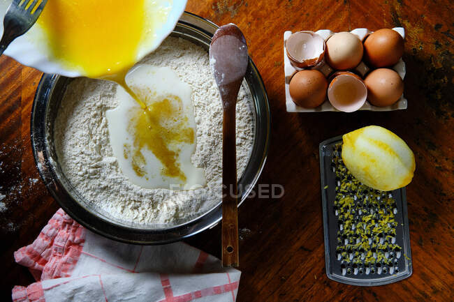 Top view of raw mixed egg spilling into bowl with flour near lemon and eggshells on table at home — Stock Photo