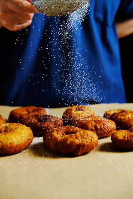 Неузнаваемый человек, использующий решето, чтобы пролить сахар-порошок на пачку свежих пончиков во время приготовления теста на кухне — стоковое фото
