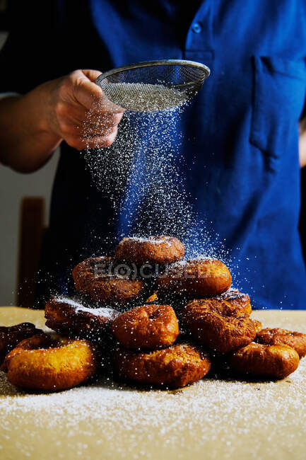 Persona irreconocible que usa tamiz para derramar azúcar en polvo en una pila de rosquillas frescas mientras cocina pastelería en la cocina - foto de stock