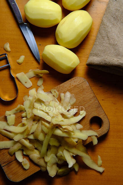 Vista superior de batatas descascadas e cascas de batata na placa de corte com faca na cozinha moderna — Fotografia de Stock