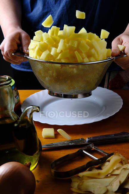 Crop female chef filtering fresh raw pieces of potatoes with sieve above white plate in kitchen — Stock Photo