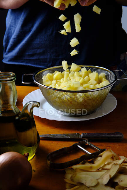 Crop chef feminino filtrando pedaços de batatas cruas frescas com peneira acima da placa branca na cozinha — Fotografia de Stock