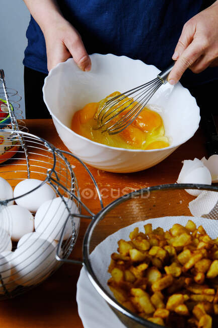 De arriba de la cosecha la mujer anónima que bate los huevos en el plato blanco sobre la mesa de madera con los ingredientes para el plato - foto de stock