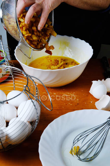 Зверху домогосподарка врожаю додає смажені скибочки картоплі до білої миски з збитими яйцями на дерев'яному столі на кухні — стокове фото