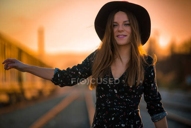 Счастливая женщина в стильной шляпе улыбается и пытается поймать транспортное средство, стоя на дороге во время захода солнца — стоковое фото