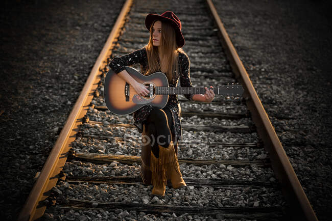 Full body stylish female playing guitar and looking away while sitting on rails during trip — Stock Photo