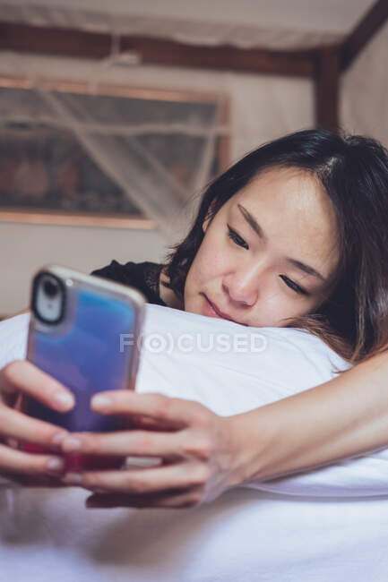 Від весела етнічна жінка посміхається і переглядає смартфон, лежачи на подушці в зручному ліжку вдома — стокове фото