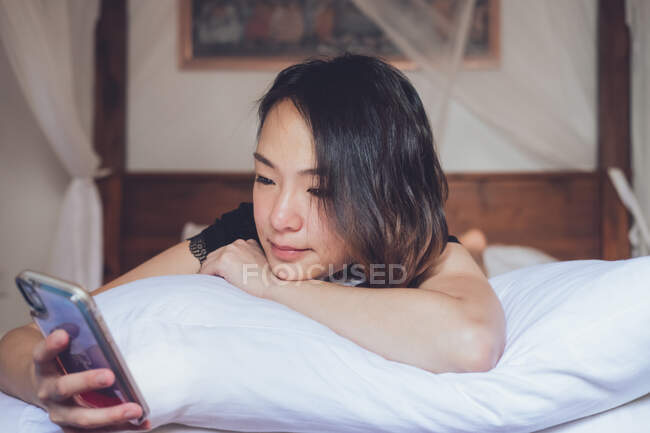 Gai sourire ethnique féminine et smartphone de navigation tout en étant couché sur l'oreiller dans un lit confortable à la maison — Photo de stock