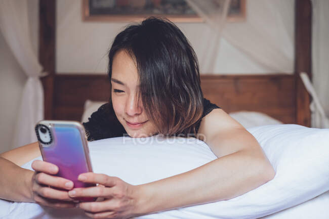Веселая этническая женщина улыбается и просматривает смартфон, лежа на подушке в удобной постели дома — стоковое фото