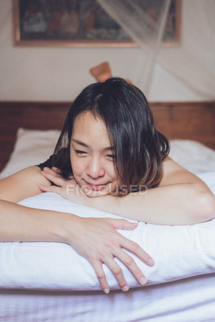 Optimistische Asiatin schaut weg, liegt auf bequemem Bett und lacht, während sie morgens zu Hause auf dem Bett liegt — Stockfoto