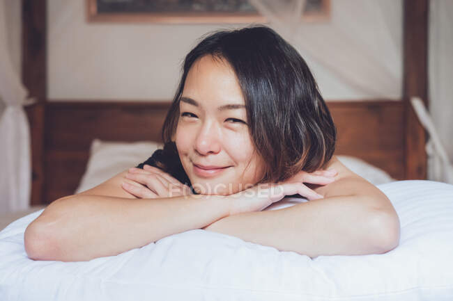 Optimistische Asiatin blickt in die Kamera, die auf einem bequemen Bett liegt und lacht, während sie morgens zu Hause auf dem Bett liegt — Stockfoto