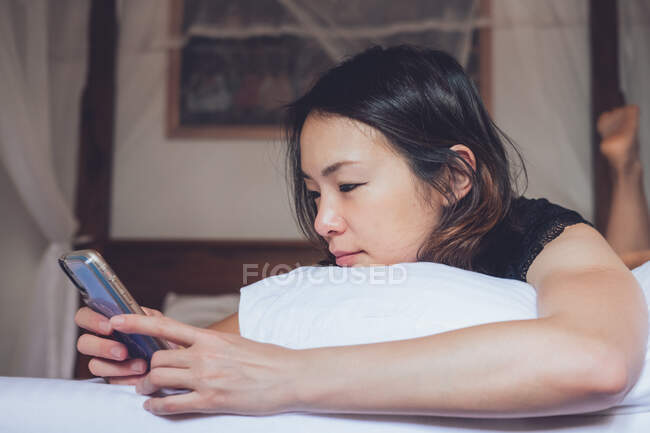 Mulher étnica alegre sorrindo e navegando smartphone enquanto deitado no travesseiro em cama confortável em casa — Fotografia de Stock